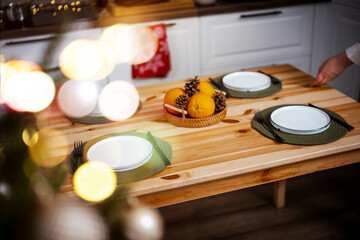 Naklejka premium Christmas wooden table setting with empty plates and fresh oranges — holiday dinner preparation. Festive home kitchen interior featuring warm fairy lights. Cozy winter evening meal anticipation.