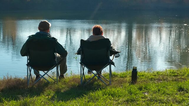 Mature couple relaxing by the lake then man leaves. Rear view of a senior couple sitting on camping chairs by a serene lake, enjoying the peaceful scenery, until the man gets up and walks away