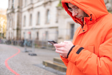 young Man in bright orange winter jacket and glasses using smartphone outdoors, male character in hooded parka texting on mobile phone, urban background with historical building, digital lifestyle © kittyfly