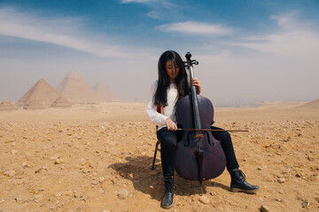 Young woman seated in focused concentration bowing a cello on the rocky sandy Giza plateau with all three Great Pyramids visible through desert haze © .shock