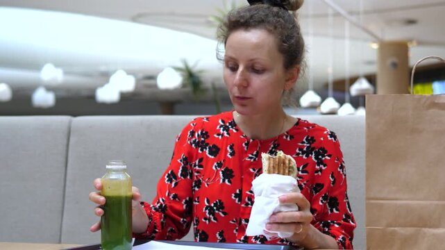 Woman lifts green juice bottle and holds wrap, young adult eating lunch in bright cafe. Woman keeps steady gaze, then lowers bottle beside paper wrapped meal in calm healthy snack setting.