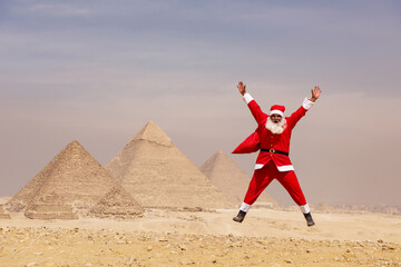 Naklejka premium Exuberant man in full Santa Claus costume leaping joyfully with arms spread wide before the iconic ancient pyramids of Giza in the Egyptian desert