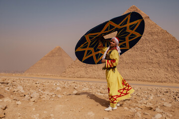 Proud folk performer in embroidered yellow galabiya and white headwrap holding bold geometric parasol at the Giza pyramids in Egypt ideal for travel © .shock