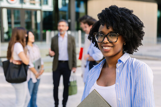 Close-up portrait of a happy young african american business school student smiling outdoors, MBA classmates blurred in background.