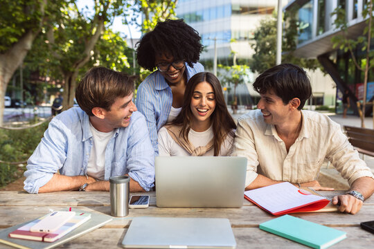 Diverse group of happy young business school students laughing while working together on a laptop outdoors. MBA classmates studying on campus. Education concept