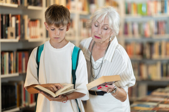 Attentive senior woman and boy choosing books near high wooden shelves in bookstore aisle