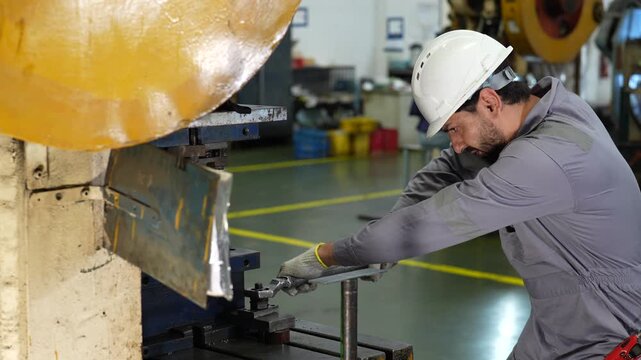 Industrial worker operating metal press machine in factory workshop, manufacturing metal production with protective helmet, gloves, gray coverall, and careful machinery handling in clean facility
