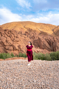 Young Latin woman smiling and walking on a wide gravel riverbed with the unique red rock formations of Dades Gorges and green trees in the background under a blue sky
