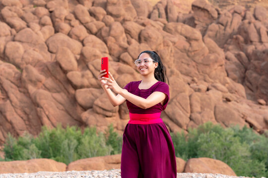 Young smiling woman wearing traditional dress and clear glasses holding a red smartphone to take a selfie in front of the unique rock formations of the Dades Valley Morocco