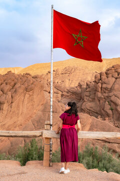 Woman standing with her back to the viewer observing the red Morocco flag waving against a backdrop of unique geological formations and a clear sky embodying travel and patriotism