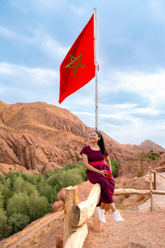 Young smiling woman enjoying the view of a desert valley with lush green oasis and mountains under a Moroccan flag symbolizing travel adventure and cultural exploration