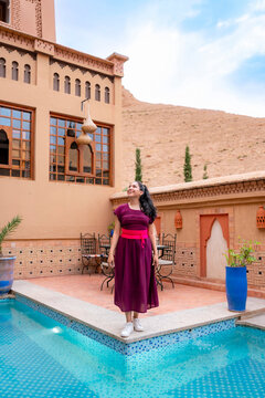 Young latin woman in a dress and sneakers standing by the pool in a traditional Moroccan riad enjoying her travel experience in Marrakech with desert hills in the background