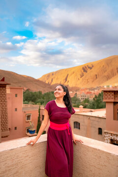 Young Latin woman smiling and looking away on a Marrakech rooftop at sunset warm terracotta tones Atlas mountains and desert skyline in the glowing evening light