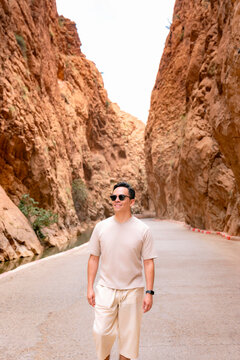 Young man smiling while walking along a sunlit road through Todra Gorge in the Atlas Mountains enjoying a solo travel adventure amid dramatic sandstone cliffs and rugged desert scenery