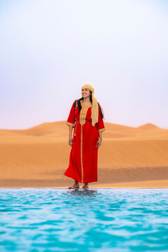 Young smiling woman wearing a red kaftan and headwrap standing barefoot at the edge of a vibrant infinity swimming pool with desert dunes in the background experiencing luxury travel