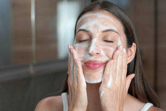 Hispanic woman washing face foaming facewash soap scrub on skin. Girl applying mousse facial cleanser for oily skin. Skin care concept.