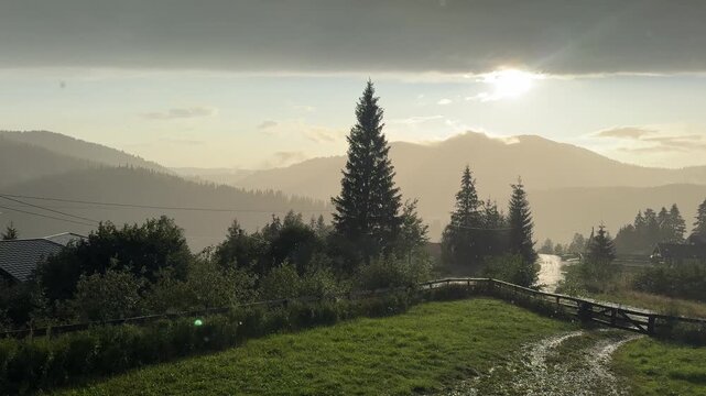 Rainfall in the Carpathian Mountains with sunlight breaking through clouds. Scenic mountain landscape, forest silhouettes and rural countryside during a summer rain shower. Peaceful nature background 