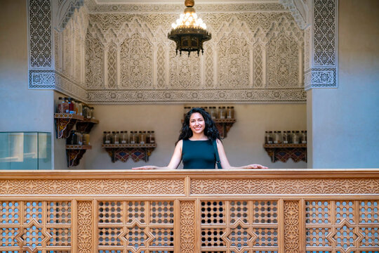 Young smiling woman standing behind a carved wooden screen enjoying a historical palace interior with traditional Moroccan decoration and spice jars on display