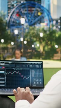 Vertical video: Man reads financial data graphs on a forex web platform using a laptop in a park, reviewing candlestick charts, market analysis trends and foreign exchange investing information.