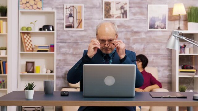 Senior man adjusts his eyeglasses at a laptop desk while a woman sits behind him, illustrating concentration, digital work and active aging through a practical home technology routine.