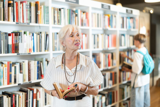 Happy senior woman choosing books near high wooden shelves in bookstore aisle