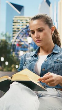 Vertical video: Casually dressed woman reads an open book and drinks coffee on a wooden bench in a park, portraying concentration, calm, mindful leisure and relaxed outdoor reading recreation time.
