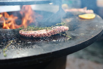 An octopus is placed on a grill over open flames. Fresh herbs and lemon slices are added for flavor. This takes place outdoors. © Anton Dios