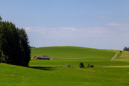 Rural Bavarian countryside landscape, Traditional wooden houses and barn on rolling hills, Lush green grass meadow against majestic Alpine mountains under blue sky and white clouds in Bavaria, Germany