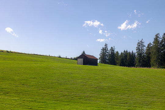 Bavarian countryside landscape, A traditional wooden barn standing alone on a rolling hill, Lush green grass meadow against majestic Alpine mountains under blue sky and white clouds, Bavaria, Germany.