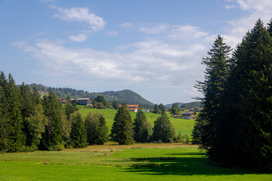 Rural Bavarian countryside landscape, Traditional wooden houses and barn on rolling hills, Lush green grass meadow against majestic Alpine mountains under blue sky and white clouds in Bavaria, Germany