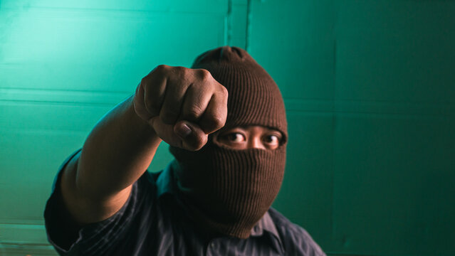 Aggressive Indonesian adult male wearing brown balaclava mask throwing punch directly at camera lens, dangerous criminal person wearing dark striped shirt isolated against green studio