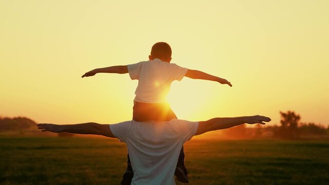 Dad plays with son, beloved child sits on father shoulders with arms raise, sun sky. Happy family plays park. Dad plays outdoors with child, dream traveling by plane. Dad child superhero fly, sunset