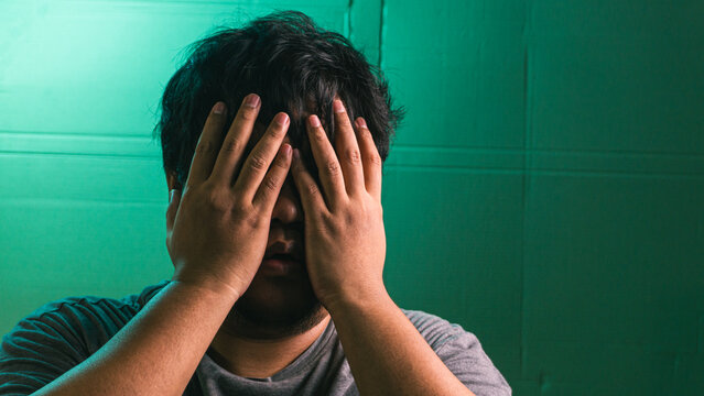 A heartbroken young indonesian male completely covers the face with both hands in a profound display of grief, shame, or exhaustion against a vibrant green textured backdrop. The hidden