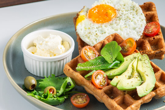 Gourmet savory waffles with avocado fried egg and cherry tomatoes on ceramic plate. Healthy breakfast or dinner platter with waffles avocado egg yolk and cream cheese