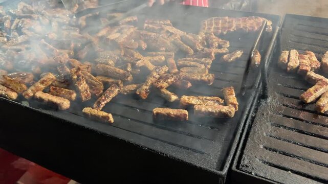 Camera pans over traditional Balkan cevapi (cevapcici) minced meat sausages sizzling and smoking on a large hot commercial grill outdoors.