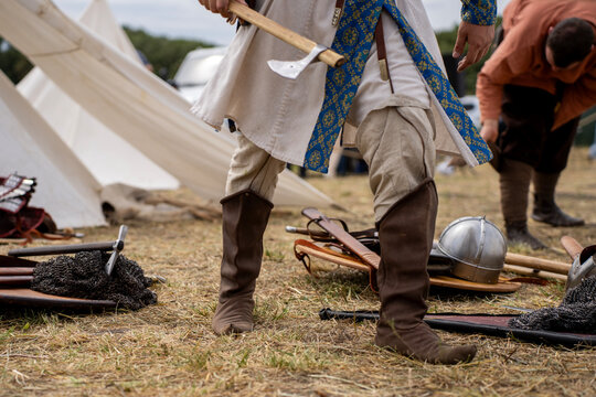 Historical reenactor in medieval tunic and leather boots holding axe standing outdoors near armor equipment and white tent camp.
