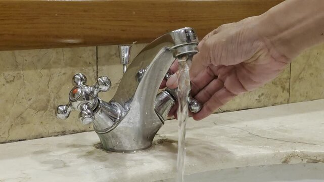 A close-up shot of a chrome double-handle bathroom faucet mounted on a marble sink countertop. A hand enters the frame to turn on the tap, letting clear water flow into the white ceramic basin, and th