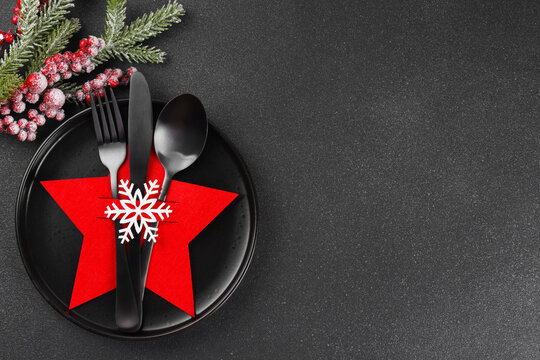 Holiday table setting with black plate and black cutlery in red star shaped cutlery holder with white snowflake and frosted Christmas tree branch on dark textured background, copy space.
