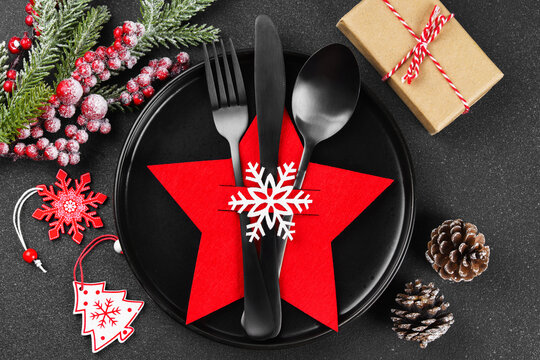 Holiday table setting with black cutlery arranged in red star shaped cutlery holder decorated with white snowflake, Christmas tree branch, ornaments and gift box on dark textured background