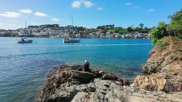 Drone footage ascends backwards revealing the picturesque town of Fowey nestled along its estuary in Cornwall. A lone individual is visible sitting on rocks by the river, appearing to take photographs