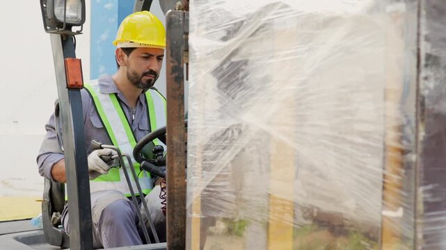 Forklift Operator Wearing Hard Hat Moves Stretch Wrap Wrapped Pallet Through Warehouse, Focused Adult Worker Handling Industrial Equipment During Safe Material Transport and Loading Operations