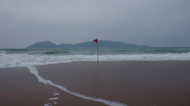 Beach with strong waves and overcast skies; swimming prohibited, marked by a red flag warning of danger. Dangerous swimming conditions marked by a red flag on a cloudy coastal beach