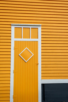 A vibrant yellow and white exterior wooden door with a triangle shaped wood window. The exterior wall is covered with narrow clapboard siding. The building foundation is concrete, painted black.