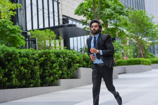 A bearded Indian businessman in formal wear sprints down the walkway with his tie loosened in front of luxury retail frontage in a central business district, in the city centre