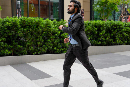 A bearded Indian businessman in formal wear races along the paving between office towers along a sunlit walkway between contemporary corporate towers, his jacket lifts behind (frame 210)