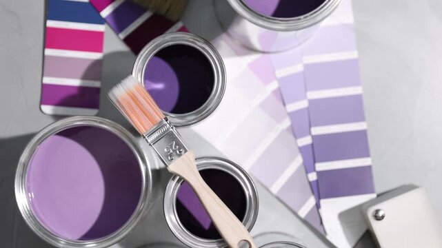 Cans with paint in shades of purple, brush and color palettes on light grey table, flat lay. Camera moving in