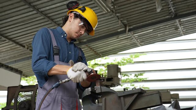 Young male industrial worker wearing helmet and protective glove, operating pneumatic tool and metal grinder beside metal vise workshop, focused craftsmanship during factory metal fabrication covered