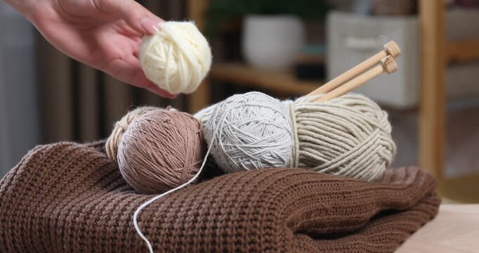 Woman putting knitting yarns onto table with sweater and needles indoors, closeup