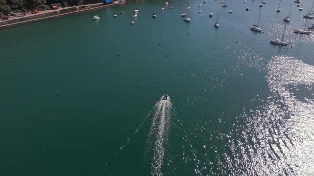An aerial perspective captures a fast rigid inflatable boat (RIB) making its way across the Fowey Estuary in Cornwall, United Kingdom. The sun glints off the water, creating sparkling reflections.