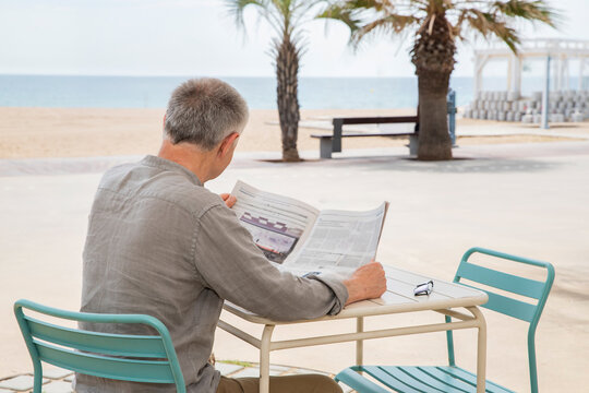 back view of senior man reading newspaper near the beach with palms